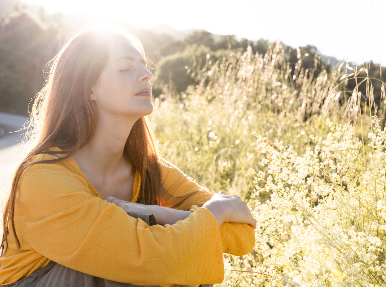 A woman in a yellow shirt is sitting in a field with her eyes closed.