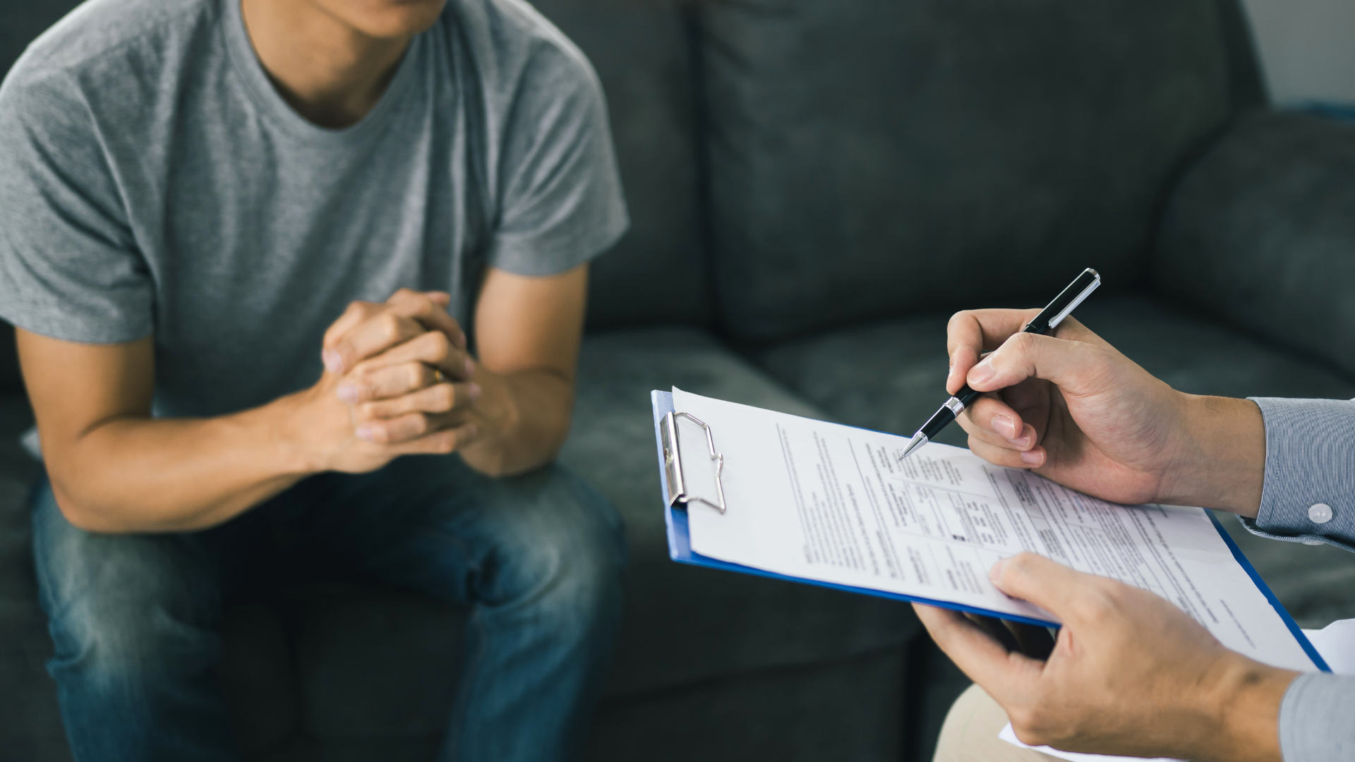 A man is sitting on a couch while a man is writing on a clipboard.