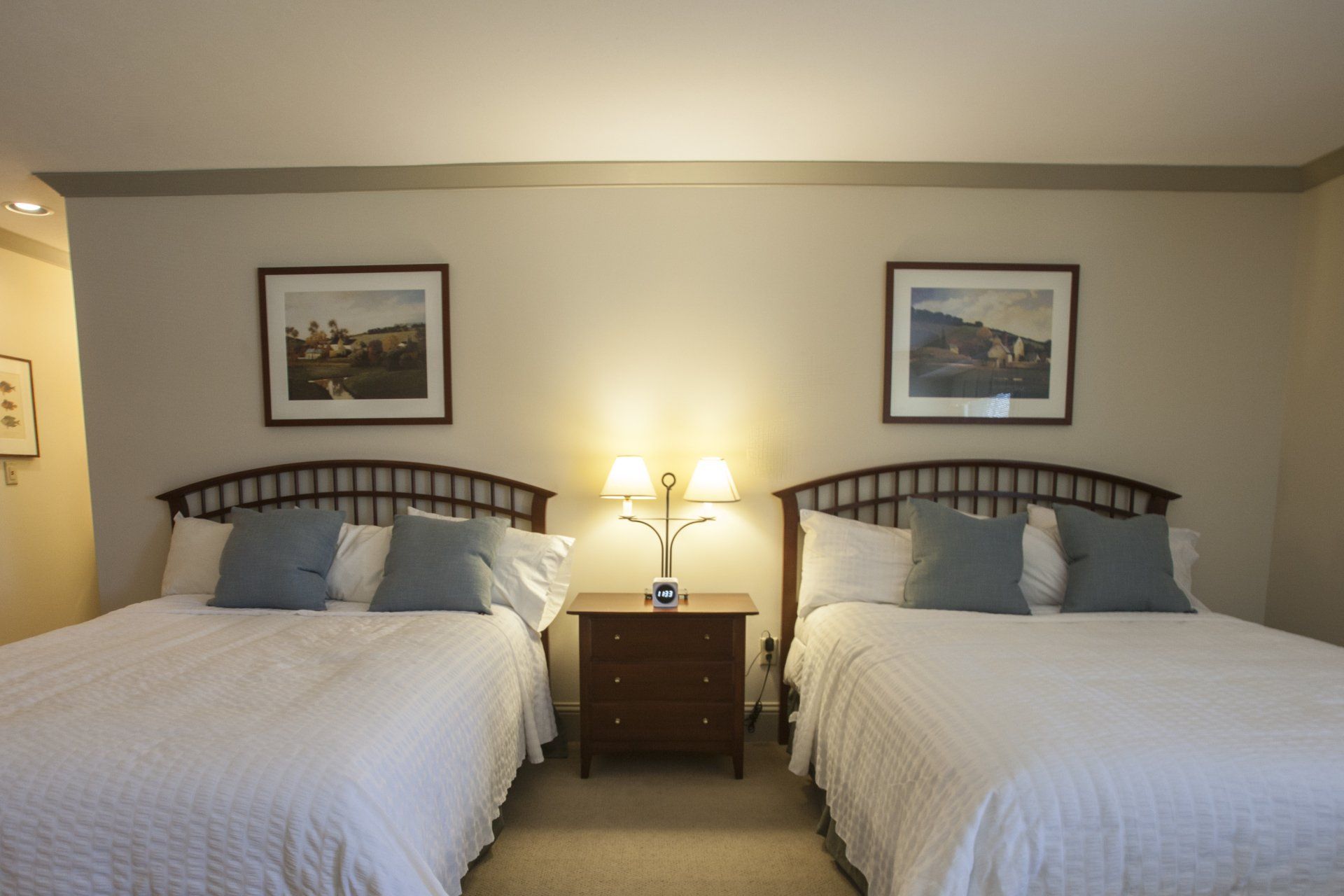 Two queen size beds with white comforter, gray throw pillows, wooden headboards and paintings above each bed.