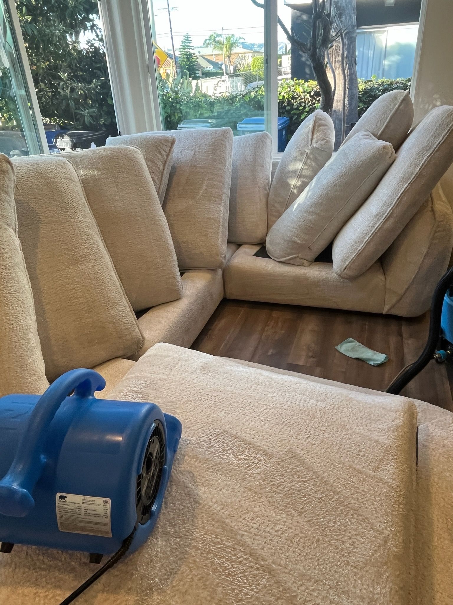 A blue industrial air mover sits on a plush, cream-colored sofa in a bright room, drying the fabric.