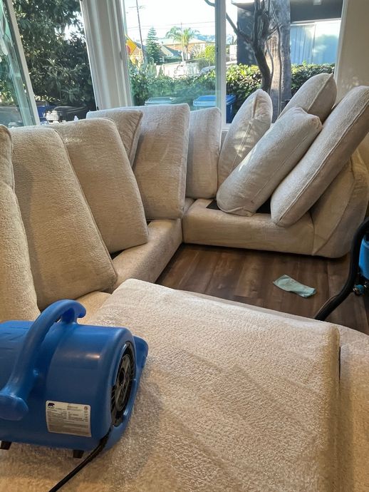 A blue industrial air mover sits on a plush, cream-colored sofa in a bright room, drying the fabric.