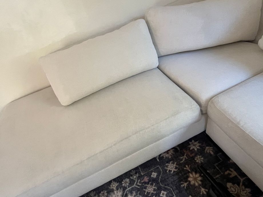 A light-colored sectional sofa with rectangular back cushions sits on a dark rug with a floral pattern.