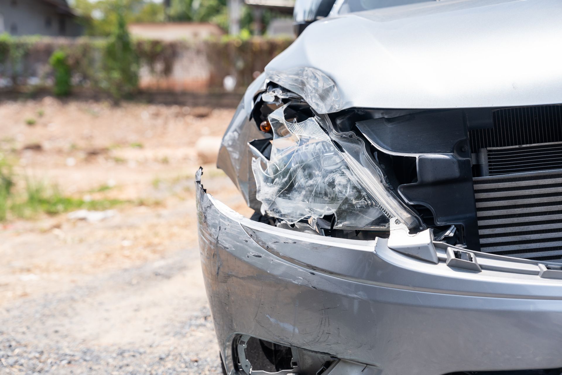 Damaged silver car front, broken headlight, dented bumper. Roadside setting.