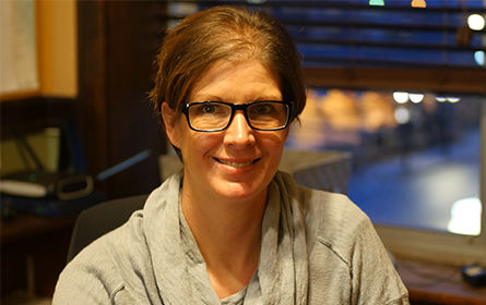 A woman wearing glasses is smiling for the camera while sitting in front of a window.