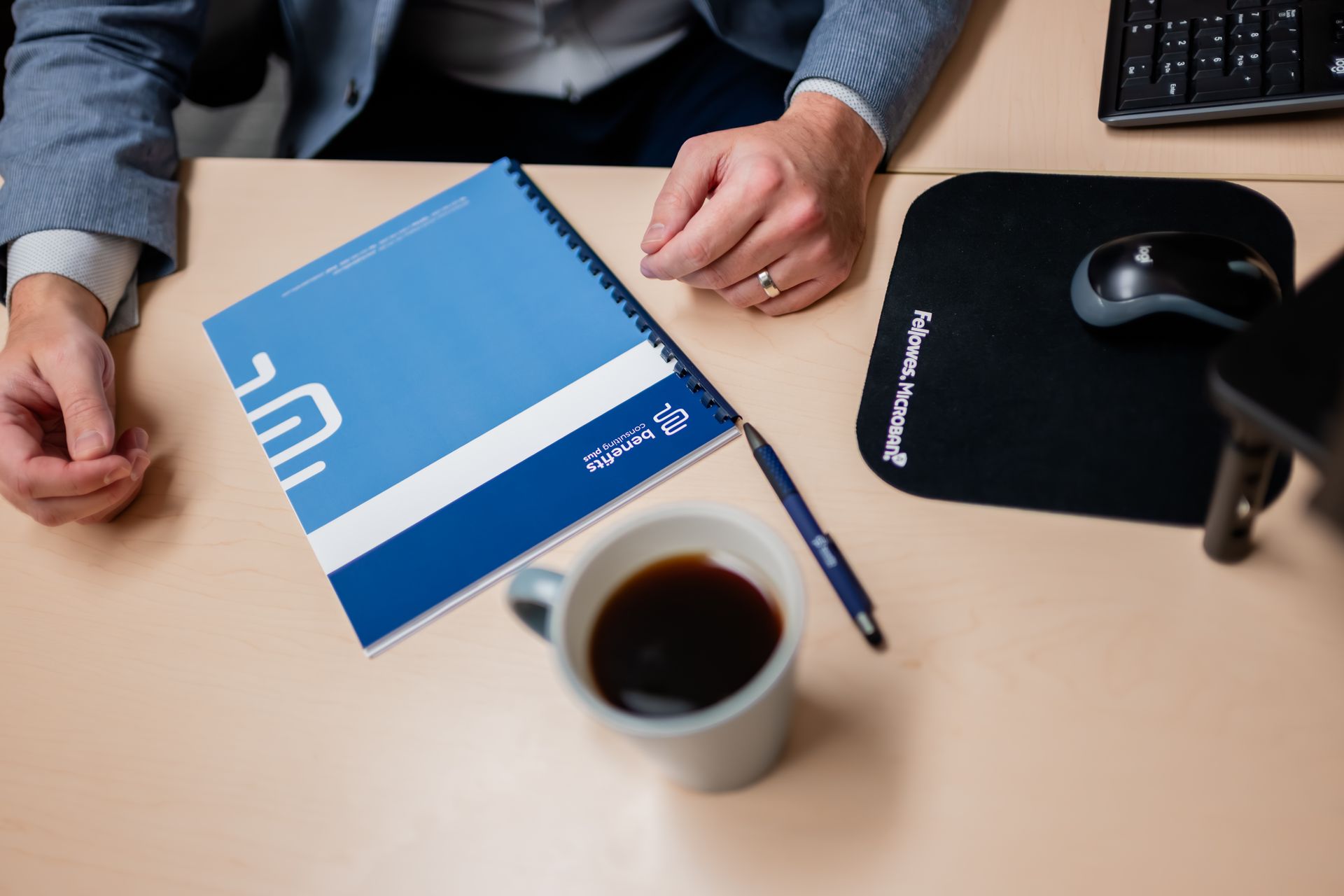 Person at desk with coffee, notebook, pen, and mousepad. Notebook is blue and white with a logo.