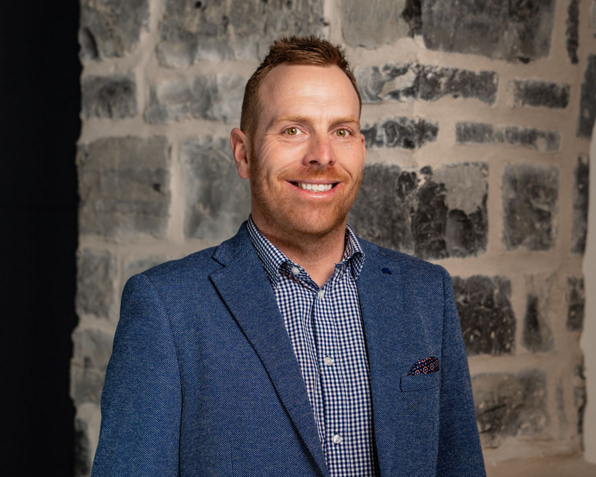 Man in blue blazer smiles in front of a stone wall.
