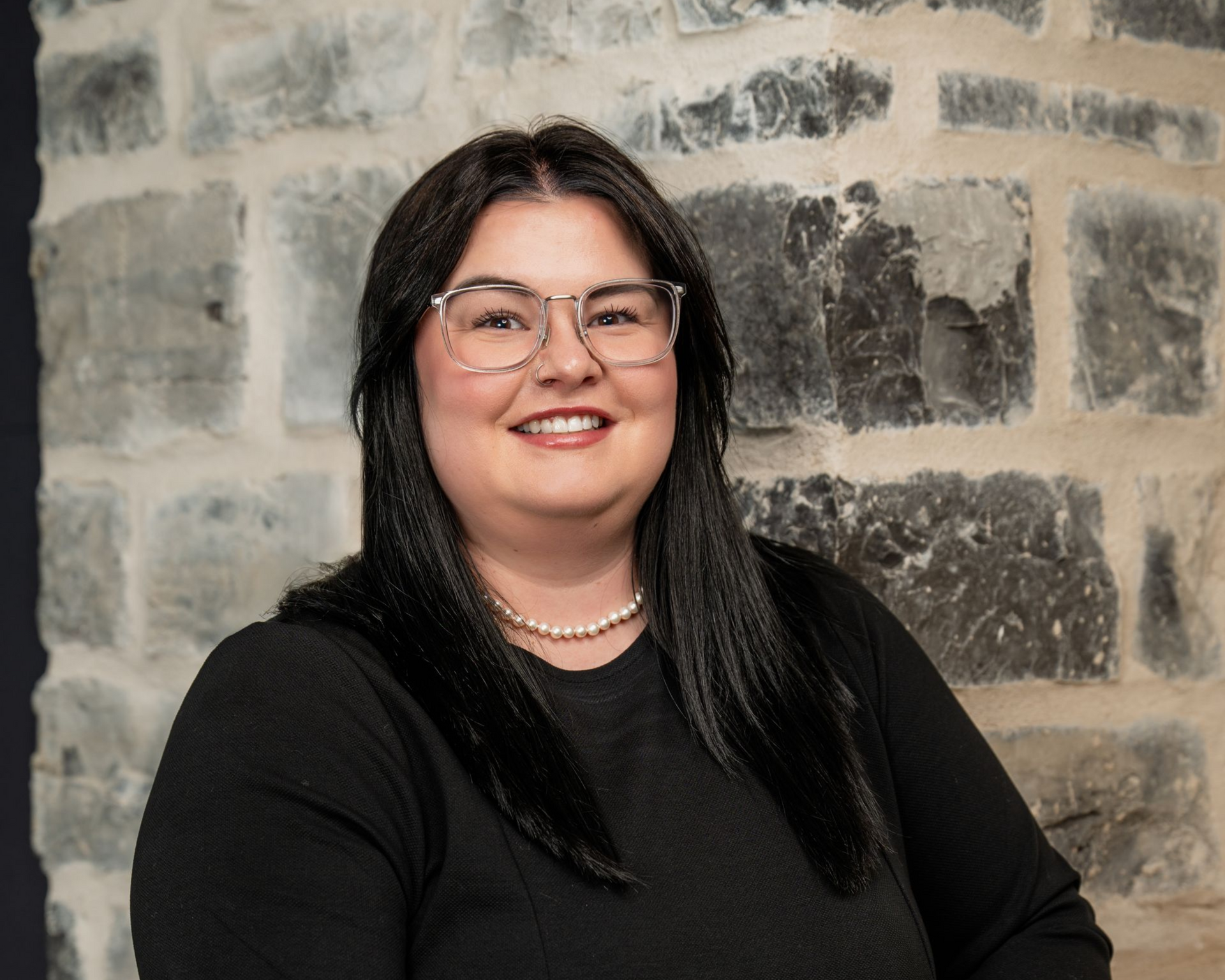 Woman with dark hair and glasses smiles in front of a stone wall.