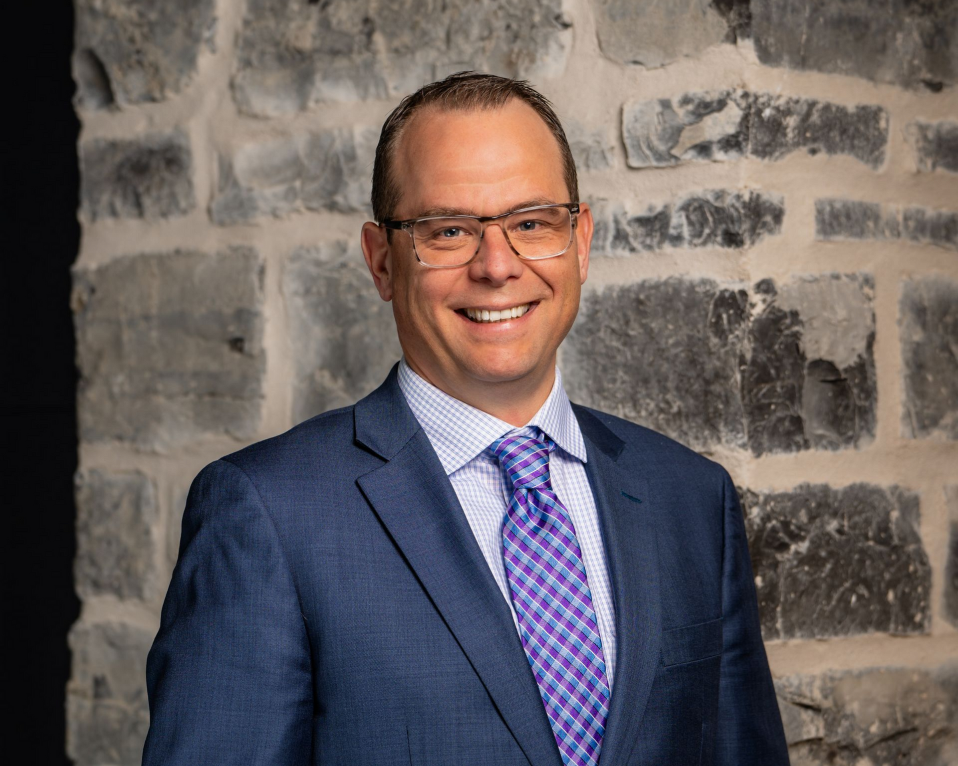 Man in blue suit, glasses, smiling, in front of a stone wall.