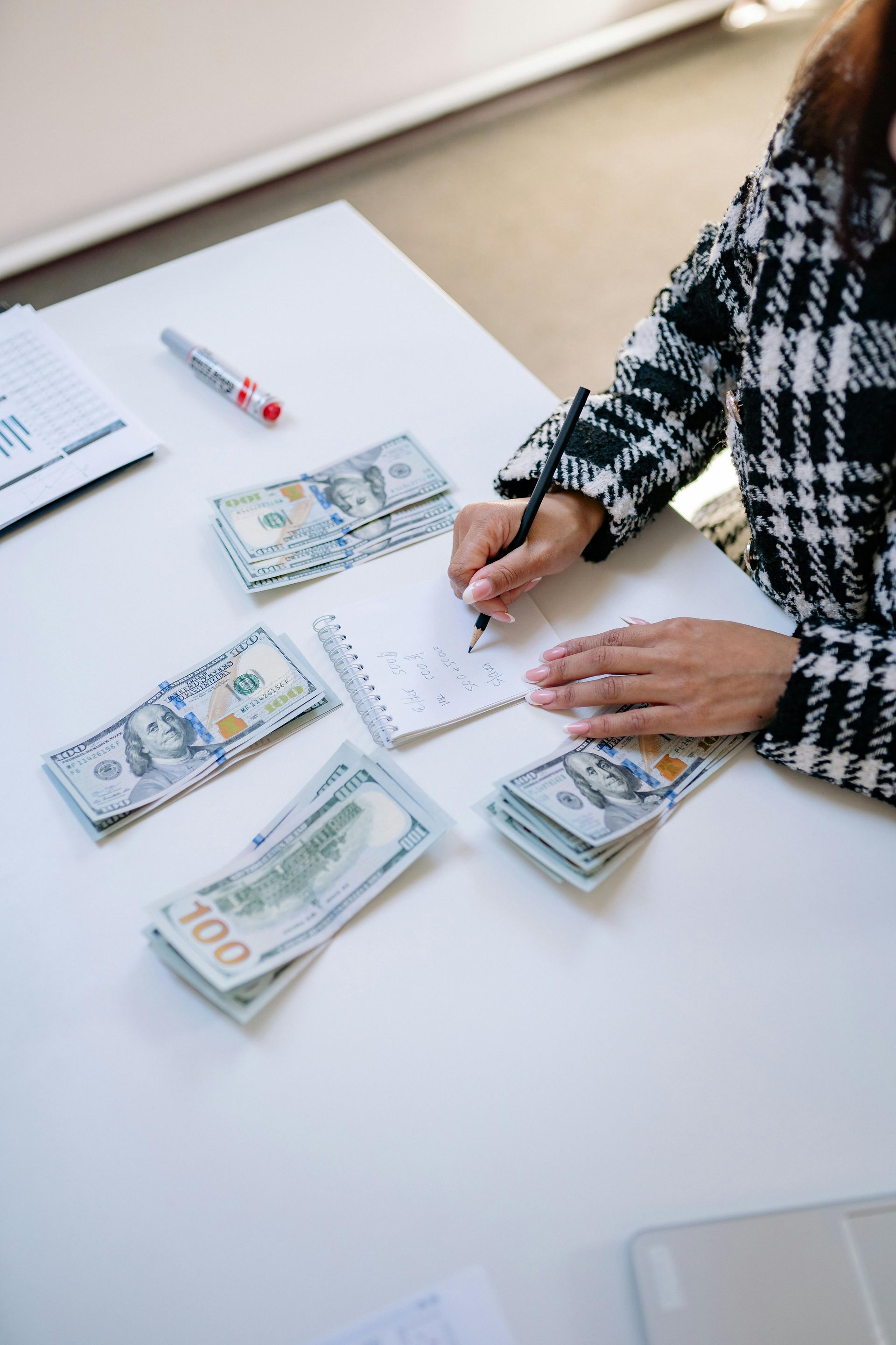 A person in a plaid jacket writes on a notepad with cash stacks on a white table.