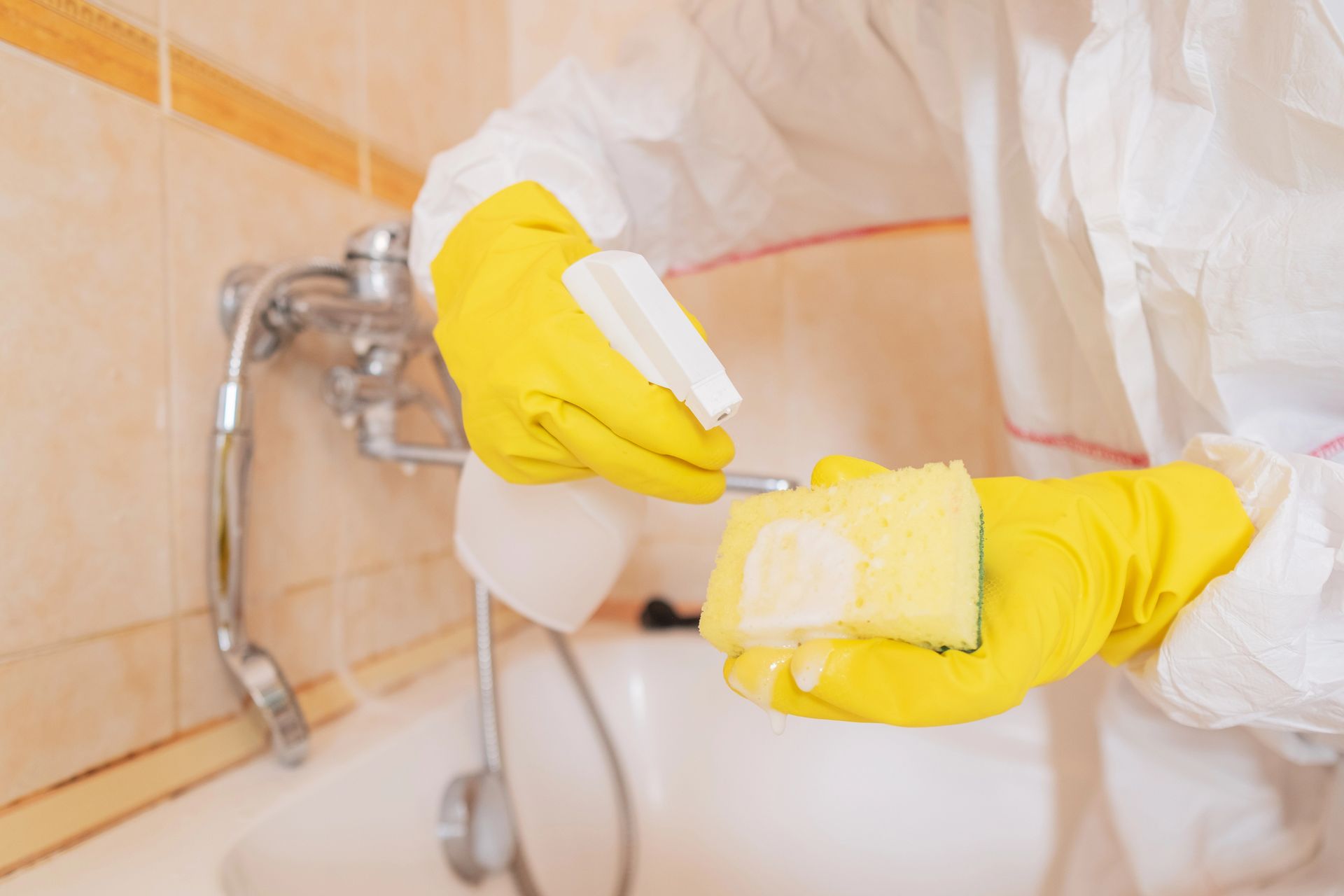 Person in yellow gloves cleaning a bathroom faucet and tile with a sponge and spray bottle