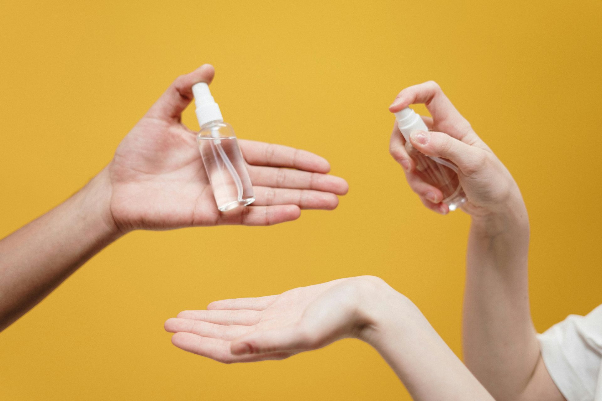 Hands spraying sanitizer onto another outstretched hand, yellow background.