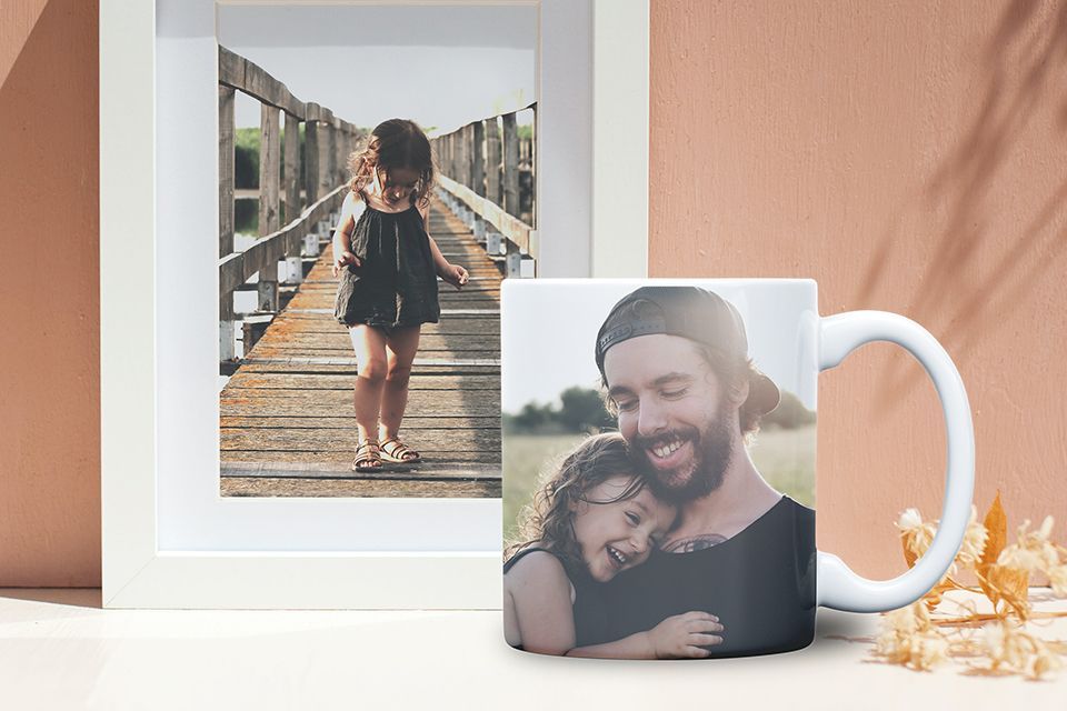 A mug with a picture of a Father and daugher printed on it  sits on a white table next to some dried flowers, in front of a white framed photo of the daughter. Both are in front of a rose colored wall.