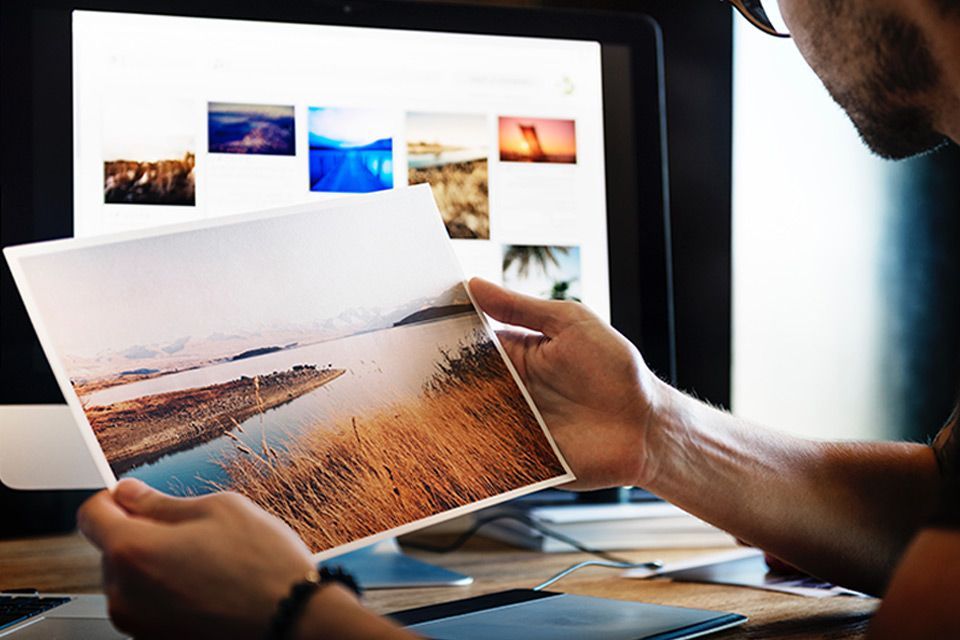 A man is holding a large printed picture  of a waterfront landscape in front of a computer screen picture photo editing software.