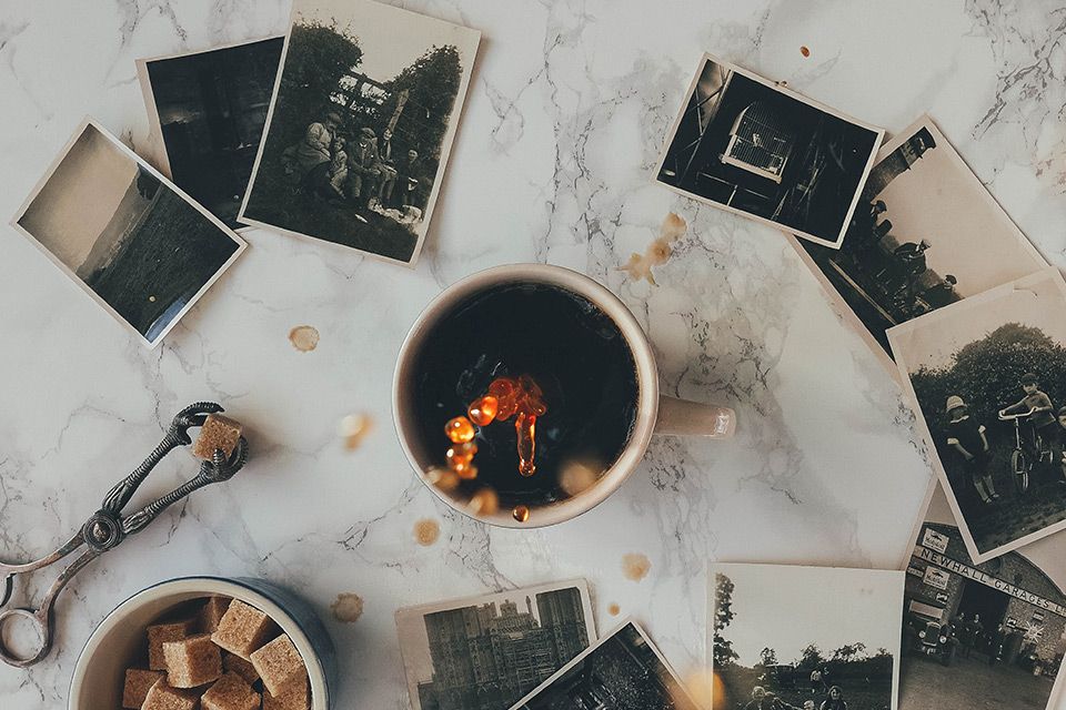 A cup of coffee is surrounded by old photographs on a marble table. It is about to spill and several drops are suspended in mid air in the shot.