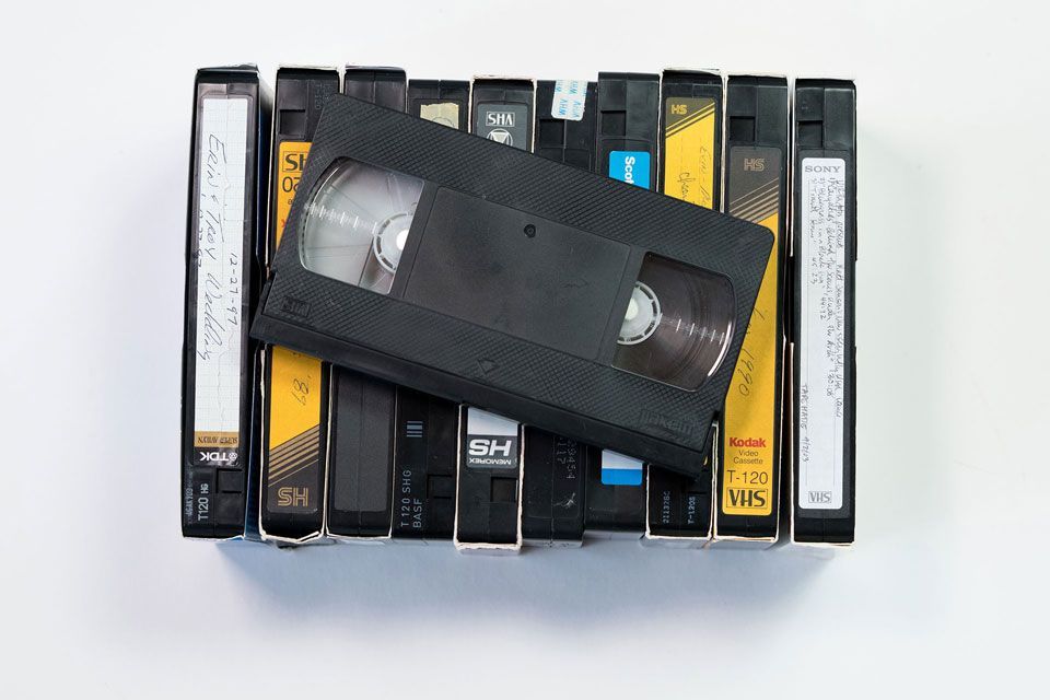 An old camera is sitting on a wooden table next to a book and a stack of photos featuring a black and white picture of a family standing next to a tree outside with houses in the background..