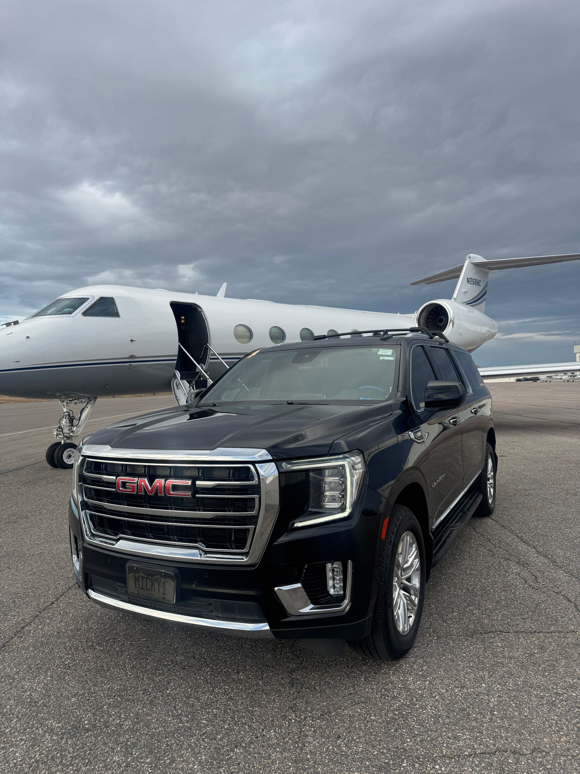 Black GMC SUV parked in front of a private jet on a tarmac under cloudy skies