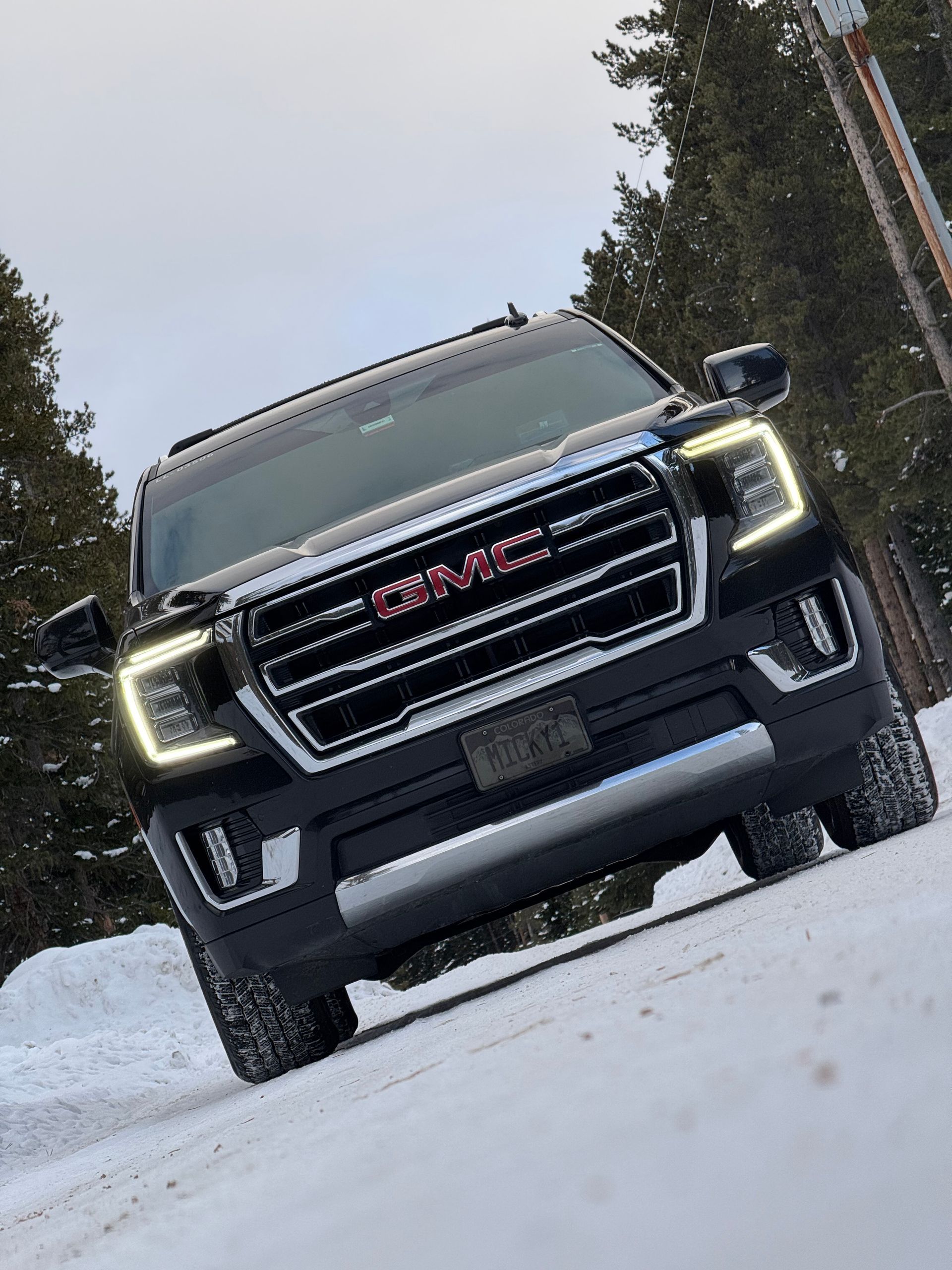Black GMC SUV driving on a snowy road, front view with headlights on and trees in the background