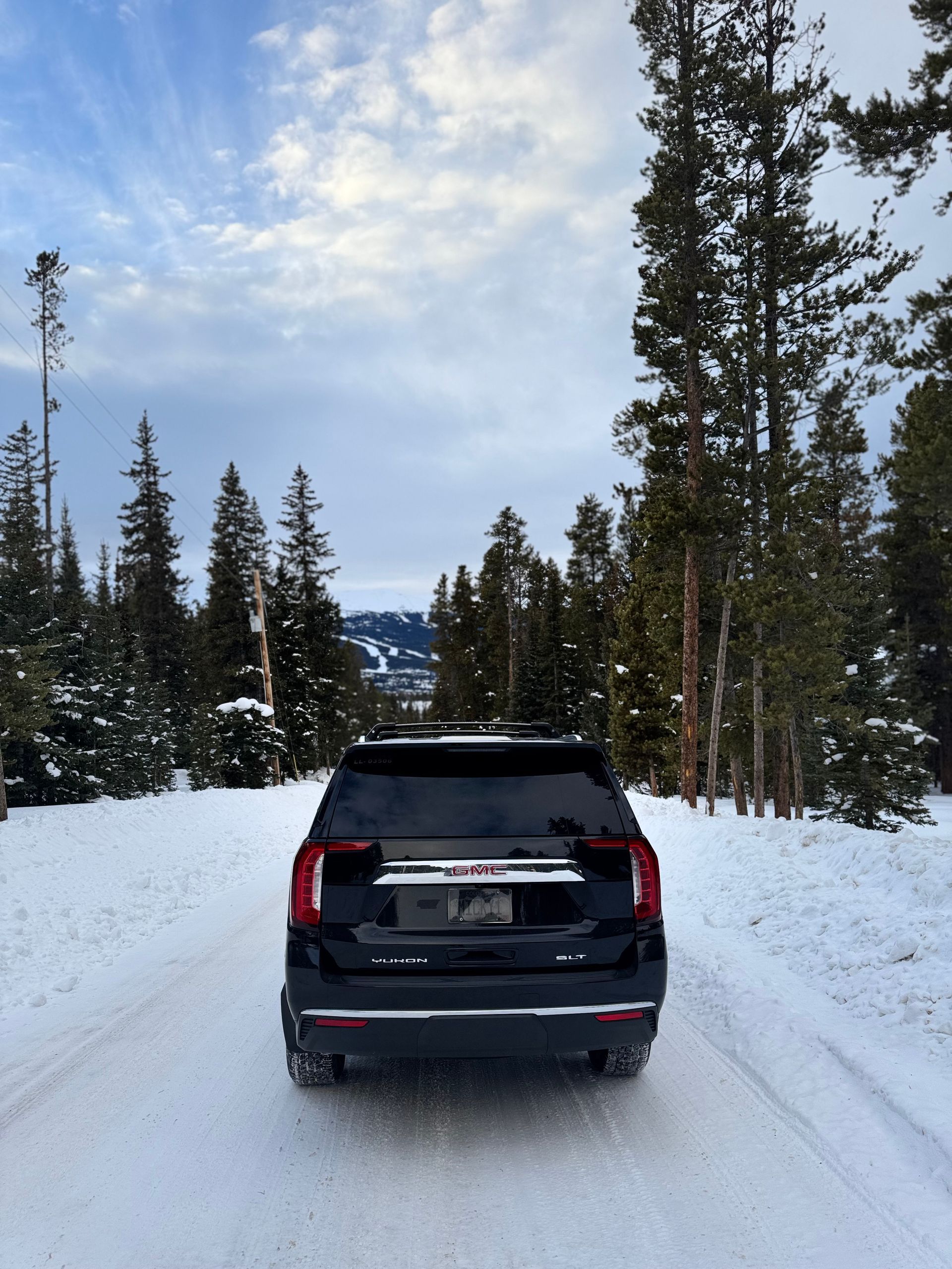 Black SUV on a snowy road lined with tall pine trees under a cloudy sky