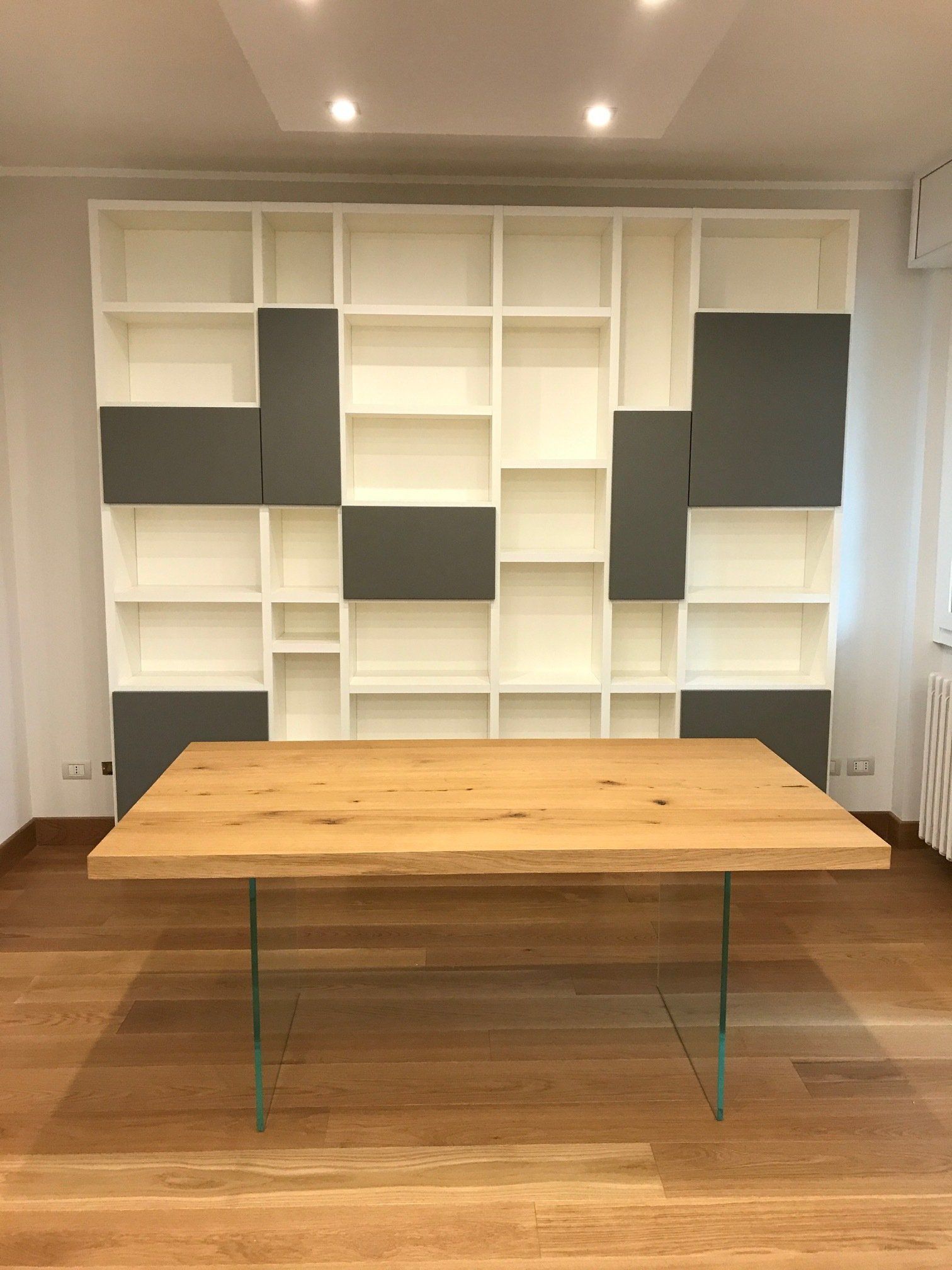 Tavolo in legno con gambe in vetro di fronte a una scaffalatura bianca e grigia.