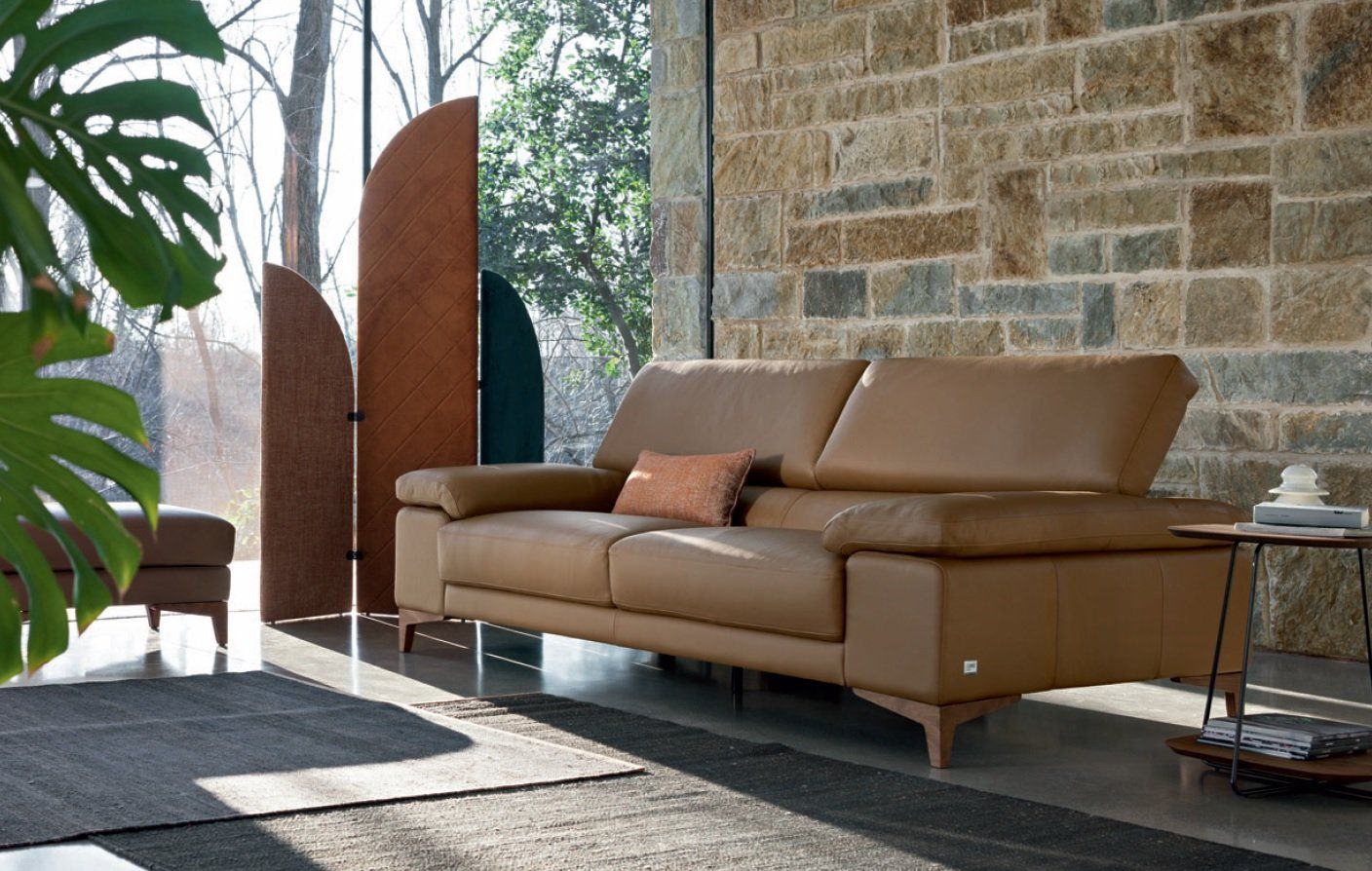 Divano in pelle in una stanza soleggiata con una parete in pietra e vista sugli alberi.