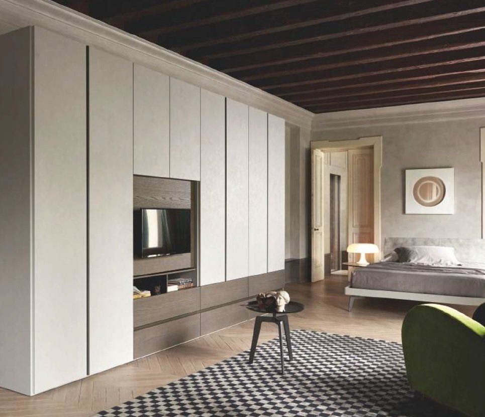 Armadio bianco a muro con TV in una camera da letto moderna, tappeto a fantasia, sedia verde, soffitto in legno.