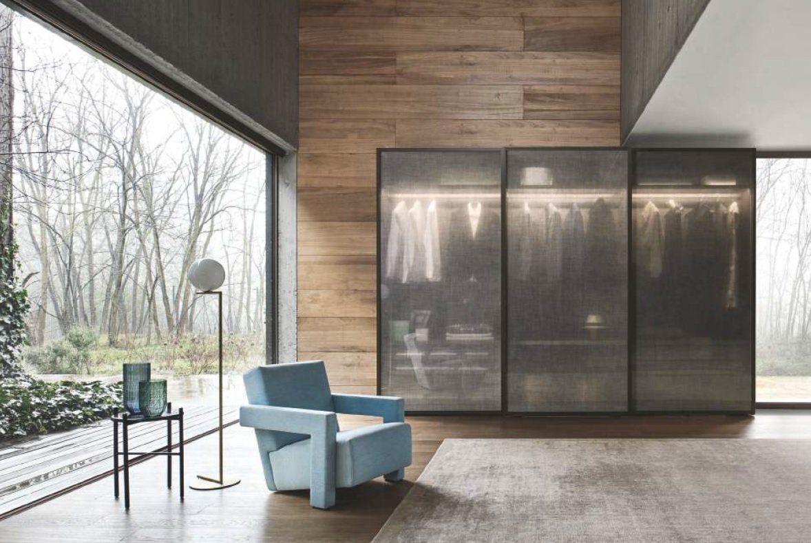 Moderna cabina armadio con ante in vetro, poltrona azzurra, lampada da terra e finestra con vista sugli alberi.