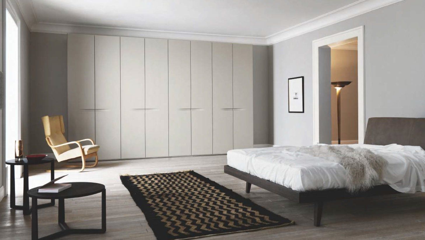 Camera da letto con un grande armadio grigio, letto, sedia e un tappeto scuro. Pareti grigio chiaro.