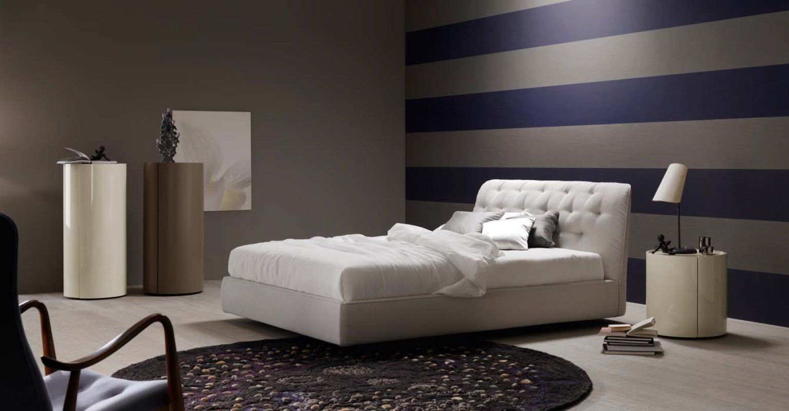 Camera da letto con letto capitonné, parete a righe blu e grigie, tappeto rotondo, tavolino e opere d'arte.