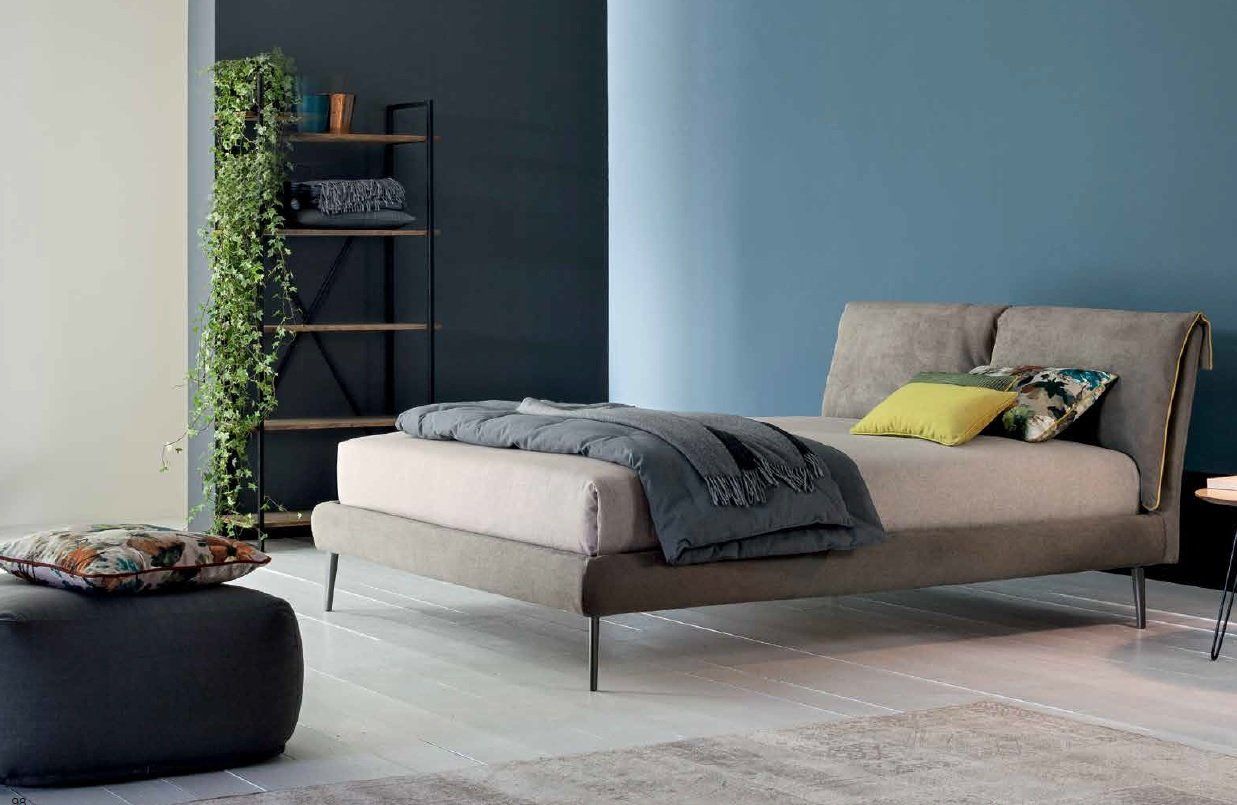 Camera da letto con letto, coperta grigia, cuscini, scaffali e una grande pianta da interno.