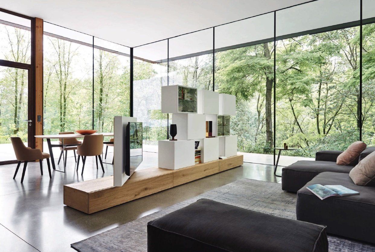 Soggiorno moderno con finestre a tutta altezza, mobili contenitori modulari bianchi, TV, zona pranzo e divano grigio.