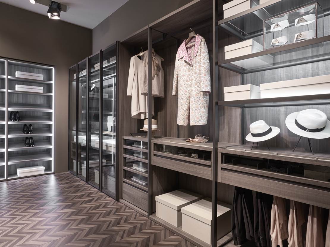 Cabina armadio con ripiani in legno scuro, ante in vetro, vestiti appesi e scatole impilate.