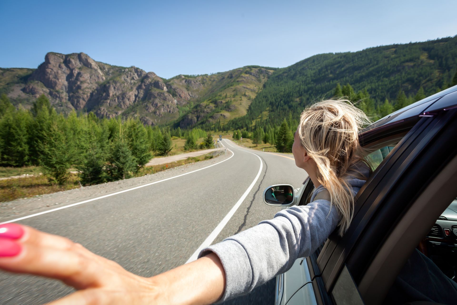 Woman leans out car window, arm extended, driving on a mountain road with greenery and peaks.