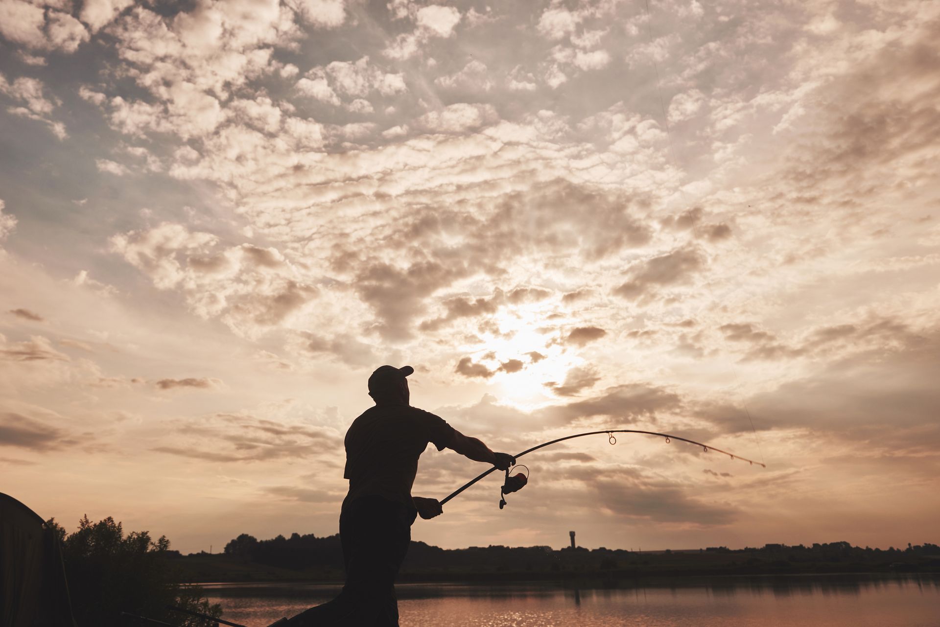 Silhouette of person fishing at sunset, casting a line into calm water. Orange and yellow sky with clouds.