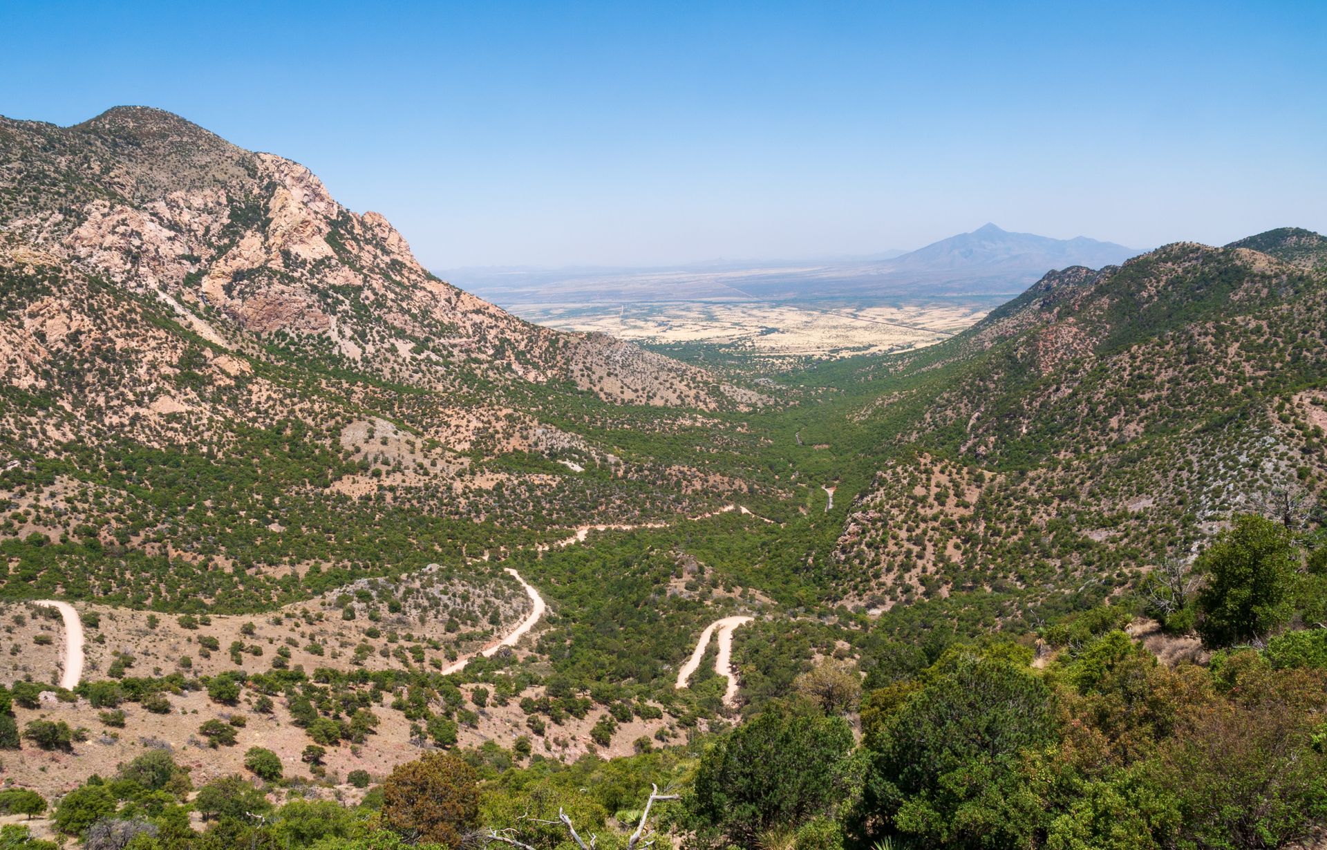 Mountain valley with winding road, green vegetation, and distant flatland under blue sky.