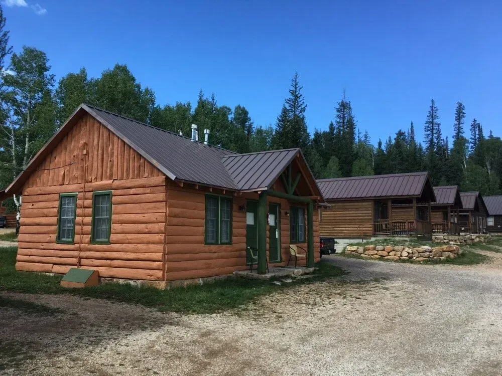 Rustic log cabins with brown metal roofs, green trim, and gravel drive under a blue sky.