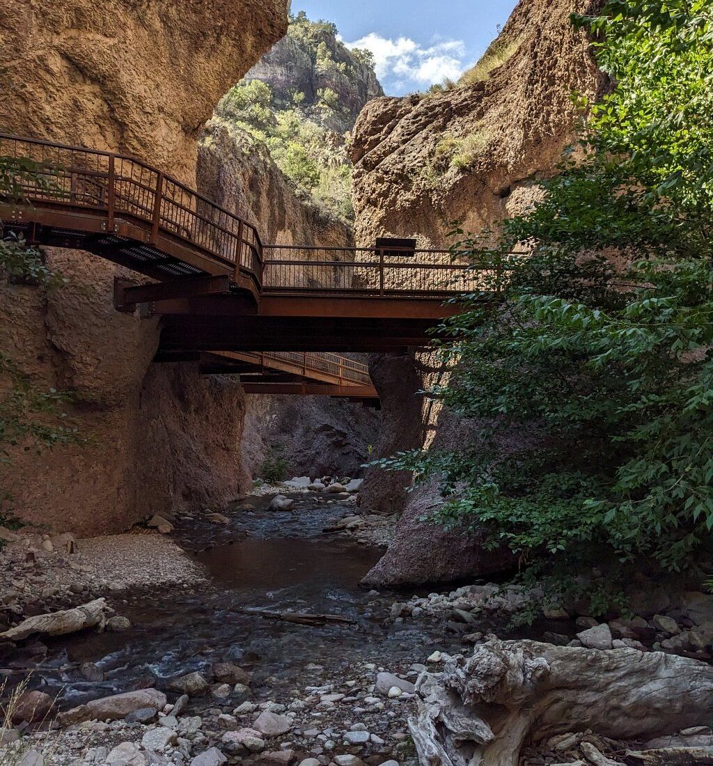 A weathered wooden walkway spans a narrow, rocky canyon over a shallow, flowing stream, surrounded by craggy cliffs.