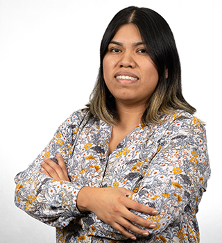 A woman in a floral shirt is standing with her arms crossed and smiling.