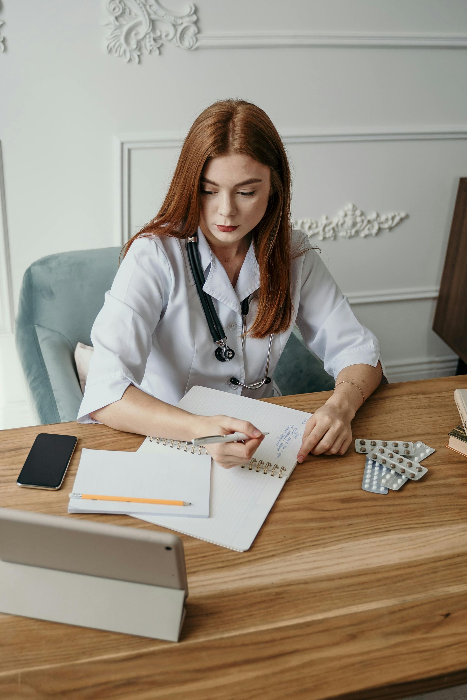 A female doctor is sitting at a desk writing in a notebook.