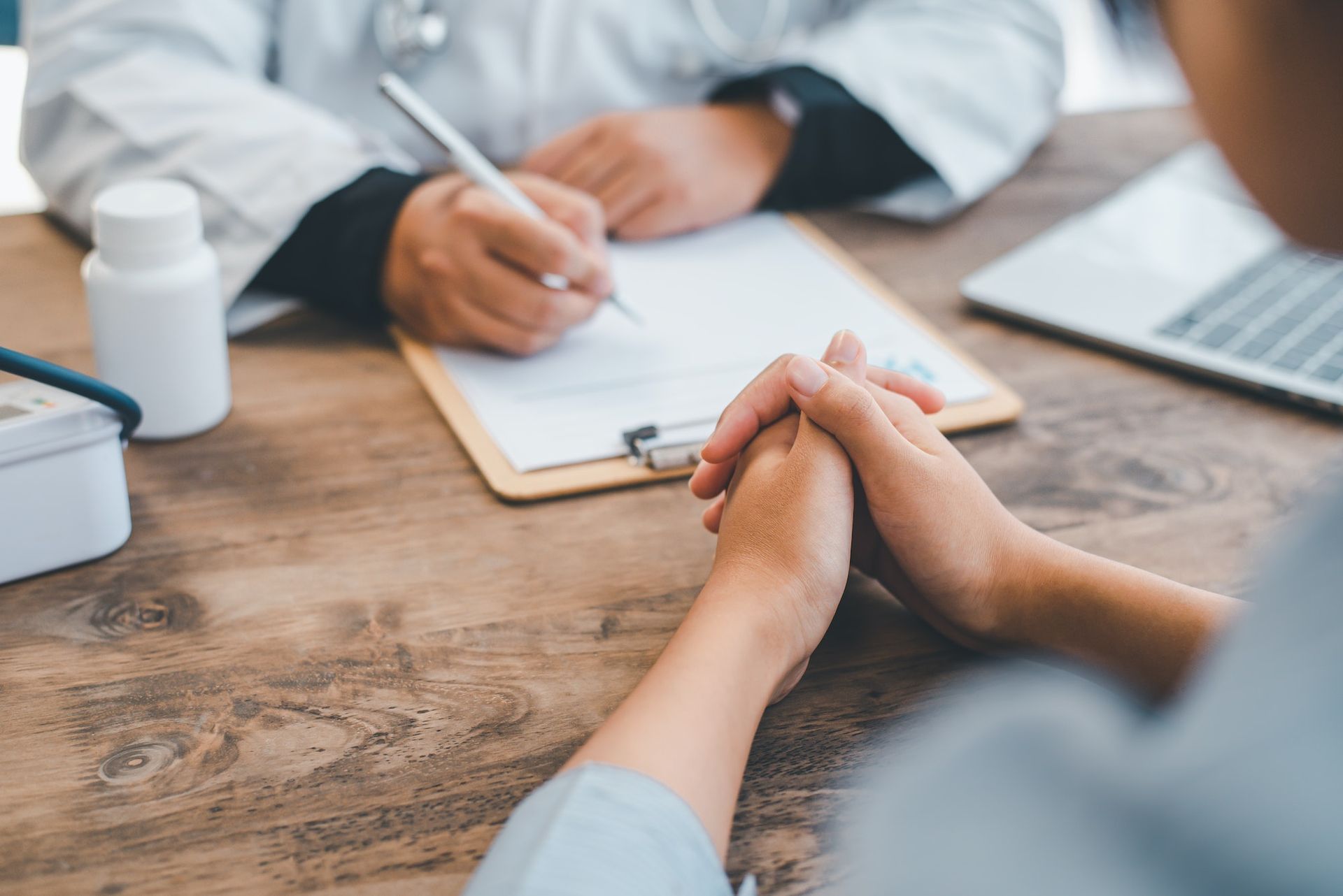 A doctor is writing on a clipboard while a patient sits at a table with their hands folded.