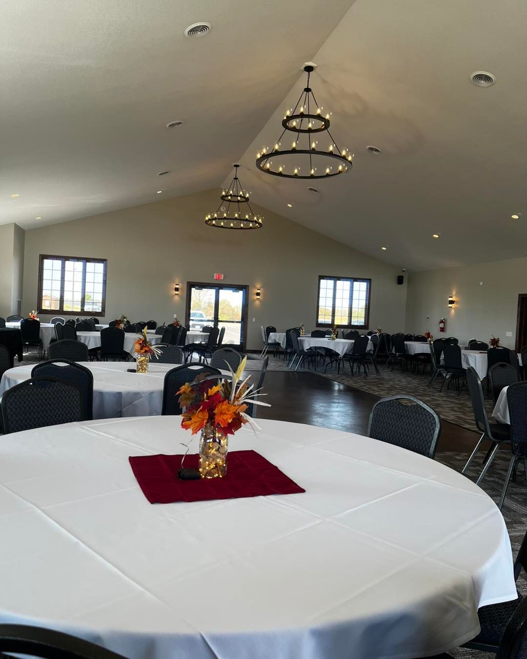 Banquet hall with round tables set for a gathering. Centerpieces of fall flowers sit on red mats.