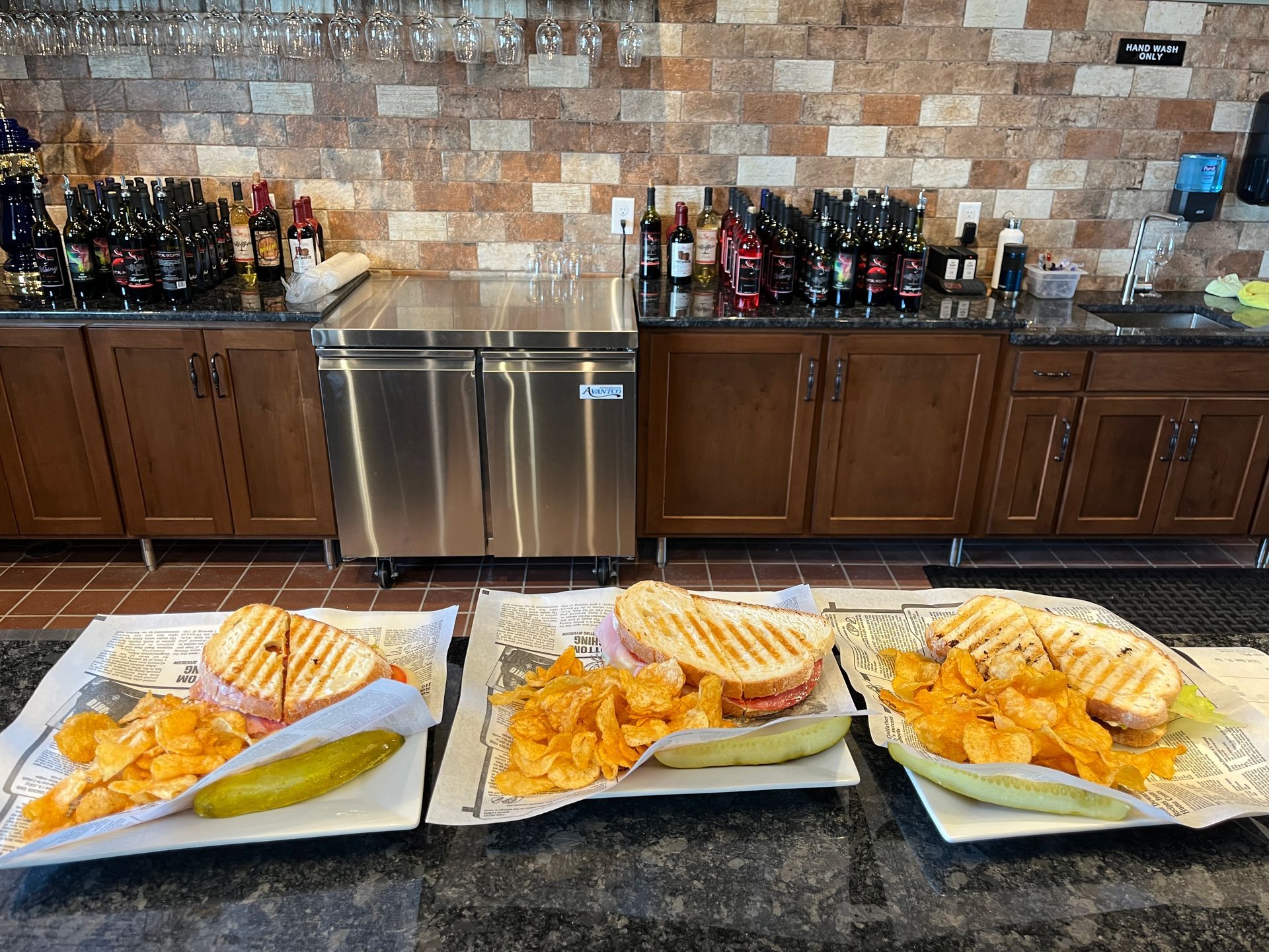Three panini sandwiches with chips and pickles on a bar, with a brick background and drinks.