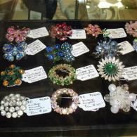 A collection of vintage brooches in various colors and designs, displayed on a black surface with price tags.