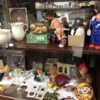 Vintage toys, including dolls, a tiger figurine, and a miniature car, displayed on two shelves in a glass cabinet.