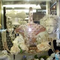A pink glass basket centerpiece surrounded by pearls, white roses, and a wedding cake topper inside a glass display case.