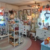A cluttered antique shop with shelves of glassware, a white chest, a glass display case, and decor items.
