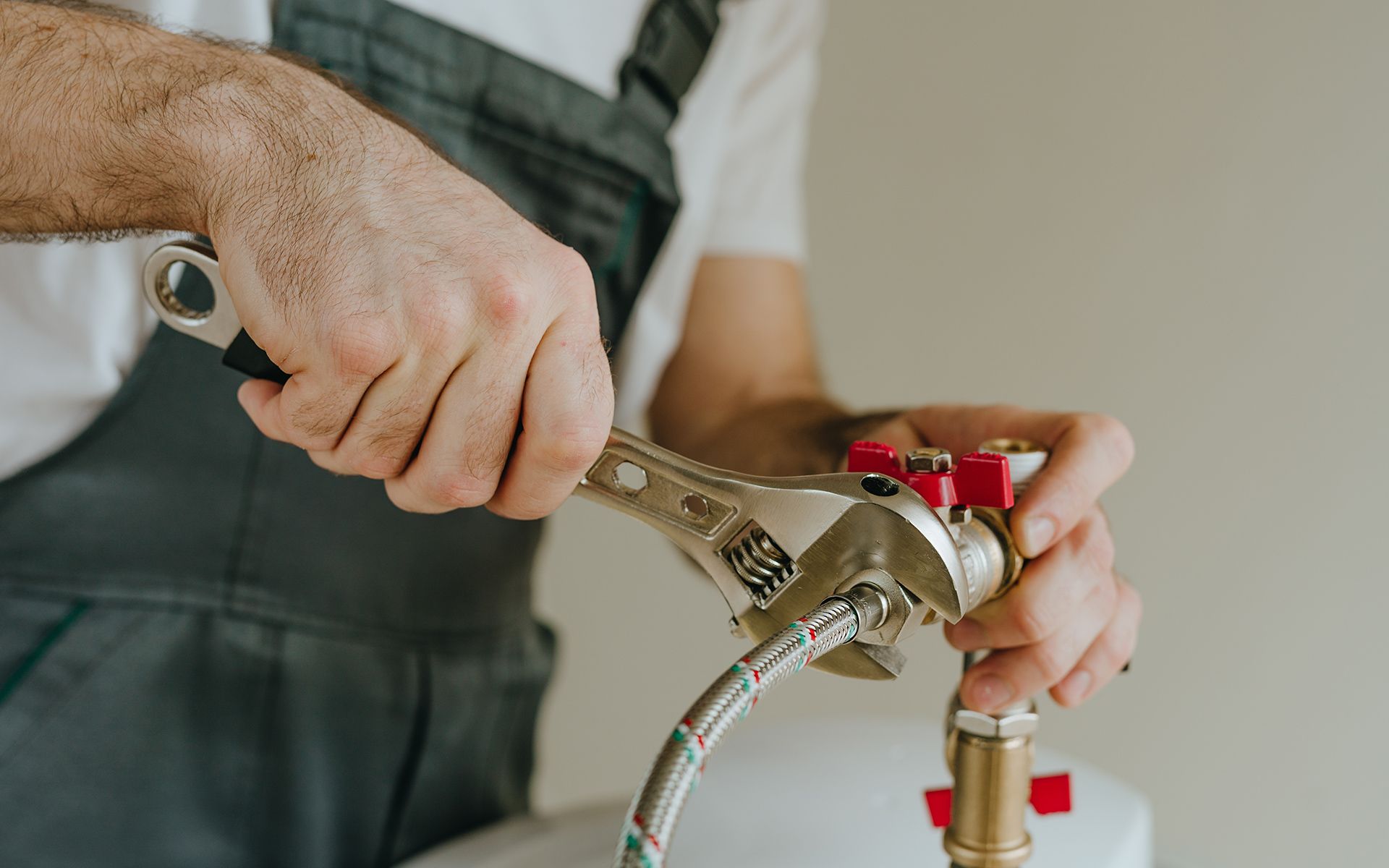 Plumber in overalls using a wrench to tighten a pipe connection near a white water heater.
