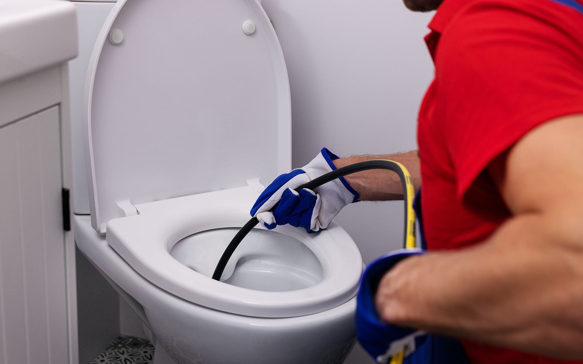 A person in a red shirt and gloves unclogs a white toilet with a snake tool.