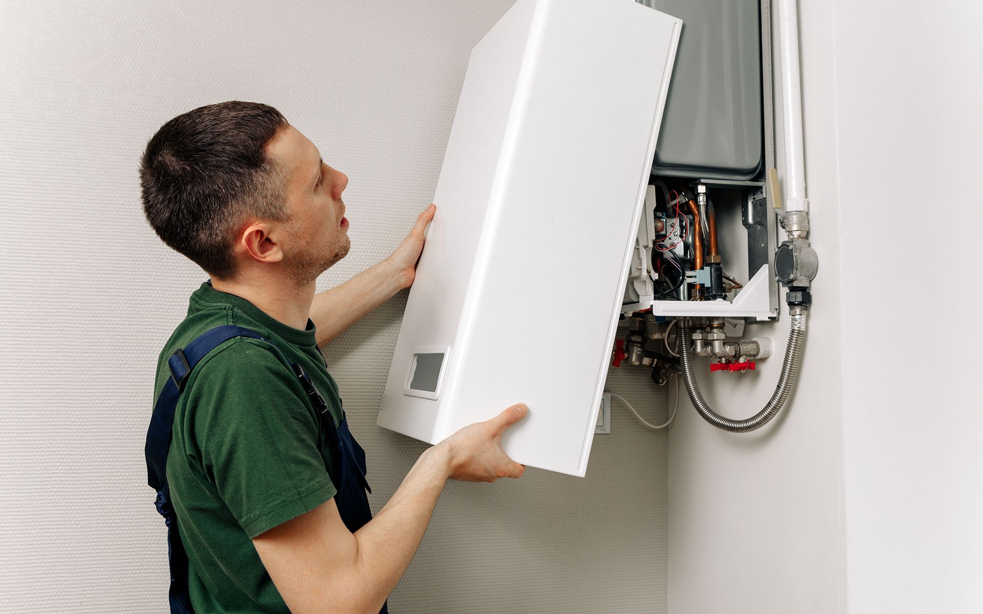 A person in overalls installing a white cover onto a heating unit on a wall.