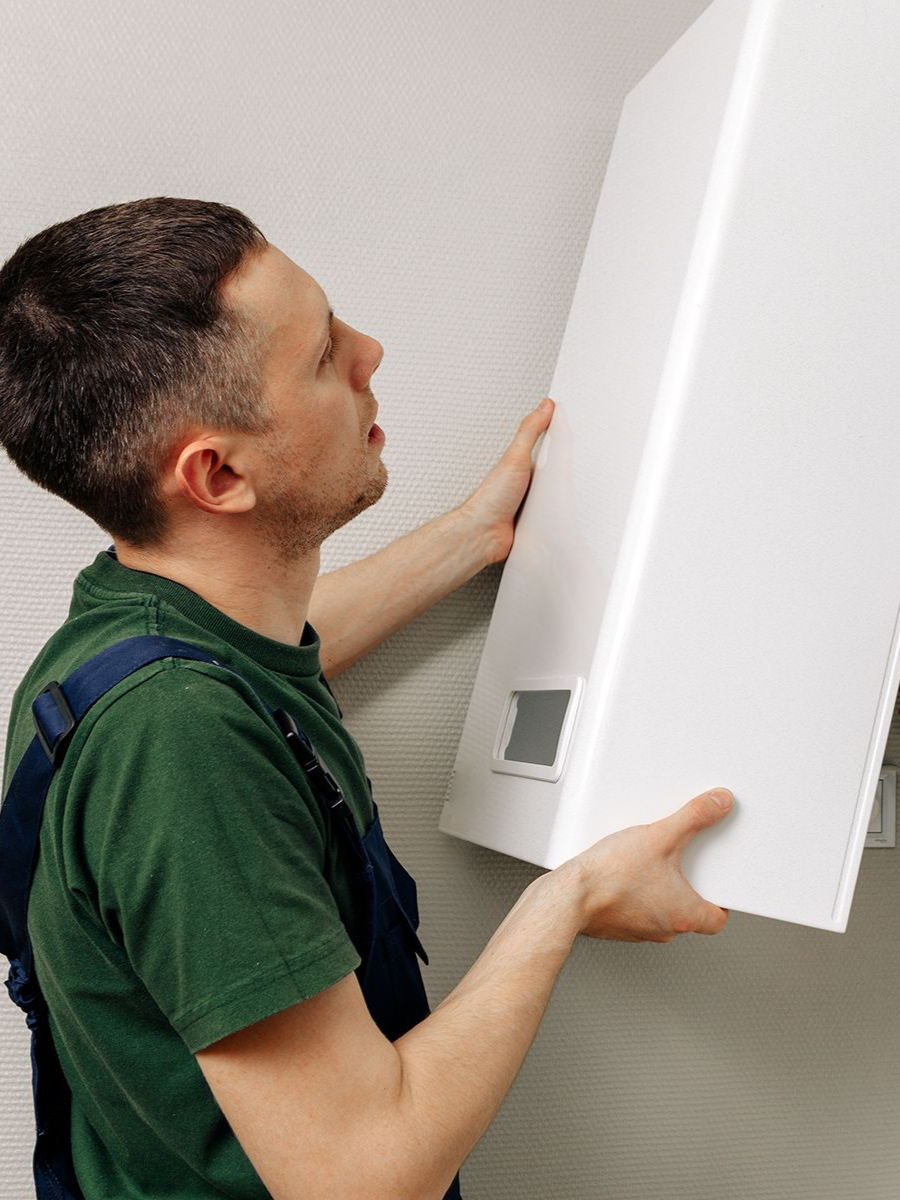 Man in green shirt and overalls installing a white appliance on a light-colored wall.