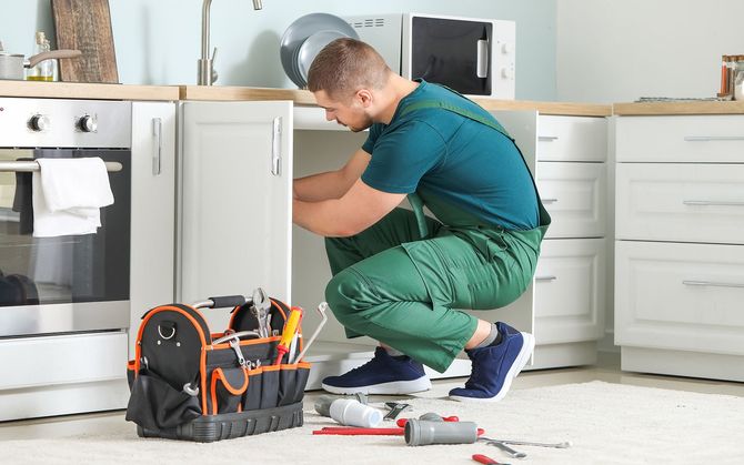 Man in green overalls working under a kitchen cabinet with a toolbox nearby.